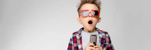 Een knappe jongen in een plaid shirt, grijs shirt en spijkerbroek staat op een grijze achtergrond. Een jongen dragen van een zonnebril. Roodharige jongen zingt in de microfoon — Stockfoto