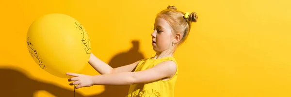 Mädchen mit roten Haaren auf gelbem Hintergrund. das Mädchen hält es in den Händen und blickt auf den gelben Luftballon. — Stockfoto