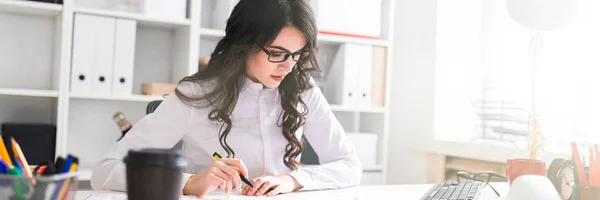 Een jong meisje zit aan de tafel van office, houdt een pen in haar hand en kijkt naar de documenten. — Stockfoto