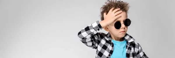 Un chico guapo con camisa a cuadros, camisa azul y pantalones vaqueros está parado sobre un fondo gris. El chico lleva gafas redondas. Niño pelirrojo sostiene su mano detrás de su cabeza — Foto de Stock