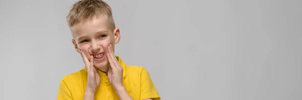Aranyos szőke kaukázusi kisfiú sárga pólót portréja ijedt kézzel közelében arca, szürke háttér — Stock Fotó