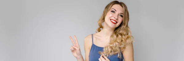Schönes Mädchen mit kräftigem Haar zeigt zwei Finger. Mädchen in einem blauen Top auf grauem Hintergrund — Stockfoto