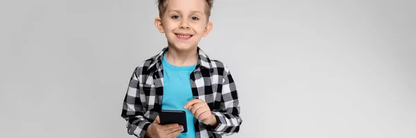 Un chico guapo con camisa a cuadros, camisa azul y pantalones vaqueros está parado sobre un fondo gris. El chico sostiene un teléfono. —  Fotos de Stock