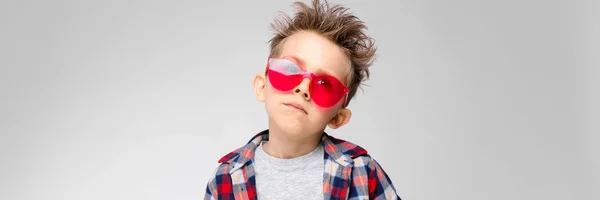Um menino bonito em uma camisa xadrez, camisa cinza e jeans fica em um fundo cinza. Menino de óculos de sol vermelhos — Fotografia de Stock