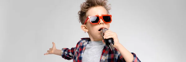 Ein gutaussehender Junge in kariertem Hemd, grauem Hemd und Jeans steht vor grauem Hintergrund. Ein Junge mit Sonnenbrille. rothaariger Junge singt ins Mikrofon — Stockfoto