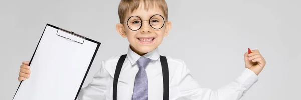 Un niño encantador con una camisa blanca, tirantes, corbata y pantalones vaqueros ligeros se levanta sobre un fondo gris. El chico sostiene un bolígrafo y sábanas para notas —  Fotos de Stock
