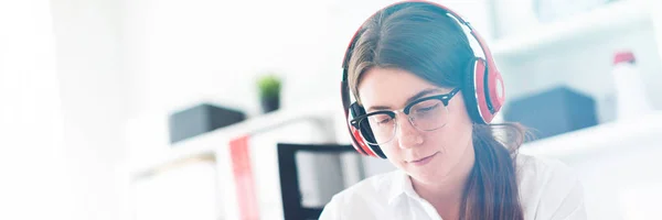 En ung flicka sitter i hörlurar vid ett bord på kontoret och innehar en telefon i handen. — Stockfoto