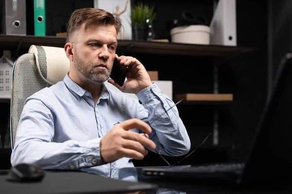 Серьезный бизнесмен, работающий над ноутбуком и разговаривающий по мобильному телефону — стоковое фото