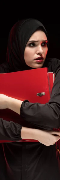 Portrait of beautiful desperate scared frightened young muslim woman wearing black hijab holding folders as keeping secret concept on black background