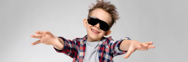 Um menino bonito em uma camisa xadrez, camisa cinza e jeans fica em um fundo cinza. O rapaz de óculos de sol pretos. O rapaz estende os braços para a frente . — Fotografia de Stock