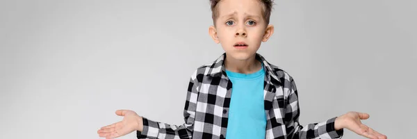 Um menino bonito em uma camisa xadrez, camisa azul e jeans fica em um fundo cinza. O menino estendeu as mãos em ambas as direções — Fotografia de Stock