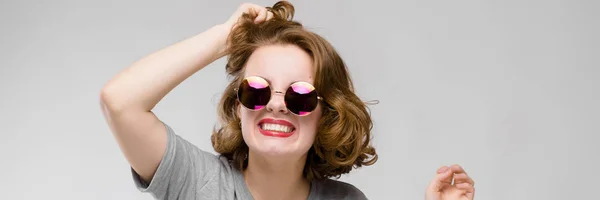 Encantadora joven con una camiseta gris sobre un fondo gris. Chica alegre en gafas redondas sostiene una mano de pelo —  Fotos de Stock