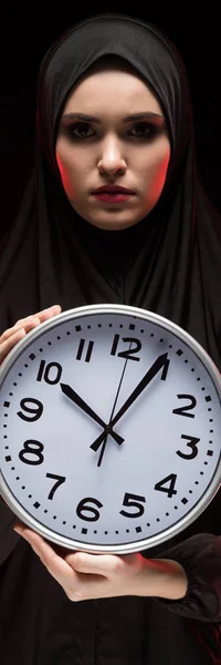 Portrait of beautiful serious scared frightened young muslim woman wearing black hijab holding clock in her hands as time running out concept on black background — Stock Photo, Image