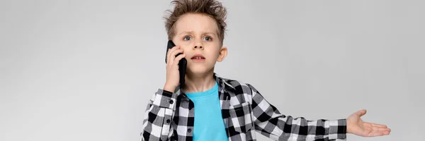 A handsome boy in a plaid shirt, blue shirt and jeans stands on a gray background. The boy is holding a phone — Stock Photo, Image