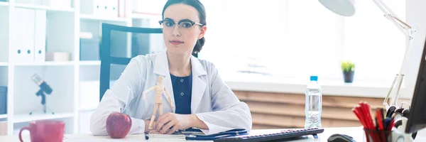 Una hermosa joven con una túnica blanca está sentada en una mesa y sosteniendo una figura de madera de un hombre en sus manos . — Foto de Stock