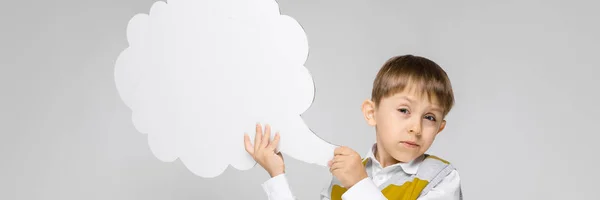 Un niño encantador con una camisa blanca, camiseta de tirantes a rayas y pantalones vaqueros ligeros está sobre un fondo gris. El niño sostiene un cartel blanco en forma de nube —  Fotos de Stock