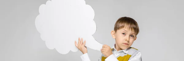 Ein charmanter Junge in weißem Hemd, gestreiftem Tank-Top und heller Jeans steht vor grauem Hintergrund. der Junge hält ein weißes Plakat in Form einer Wolke — Stockfoto