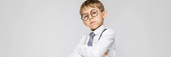 Um menino encantador com uma camisa branca, suspensórios, uma gravata e jeans leves fica em um fundo cinza. O menino dobrou os braços sobre o peito — Fotografia de Stock