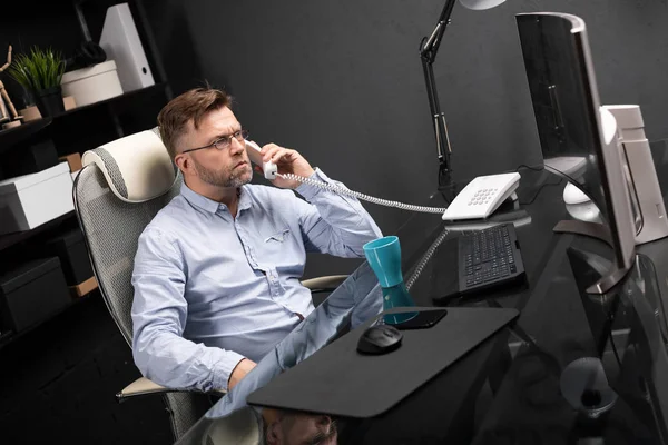 Серьезный бизнесмен работает в офисе за компьютерным столом и разговаривает по стационарному телефону — стоковое фото