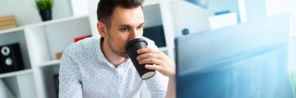 A young man stands near a table in the office, holds a pencil in his hand and drinks coffee. A young man works with documents and a computer.