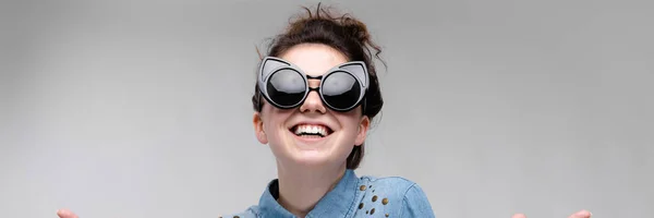 Junges brünettes Mädchen mit schwarzer Brille. Katzenbrille. Die Haare sind zu einem Dutt zusammengebunden. das Mädchen zieht ihre Handflächen nach vorne. — Stockfoto