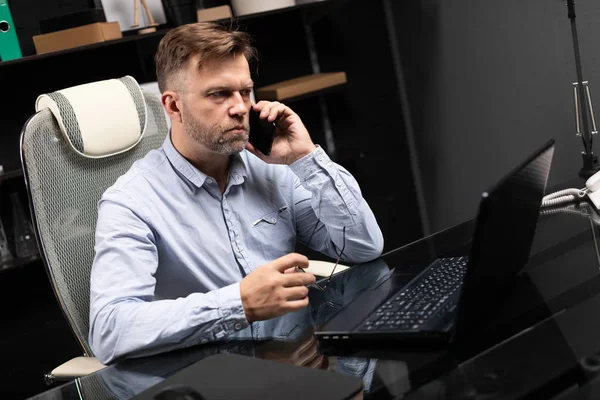 Серьезный бизнесмен, работающий над ноутбуком и разговаривающий по мобильному телефону — стоковое фото