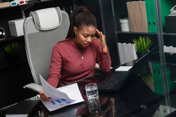 Omtänksamma unga afroamerikansk affärskvinna hålla händerna bakom huvudet och arbeta med dokument på skrivbord i office — Stockfoto