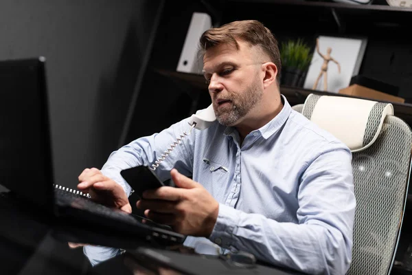 Молодой человек сидит за компьютером стол и держит стационарный телефон и мобильный телефон — стоковое фото
