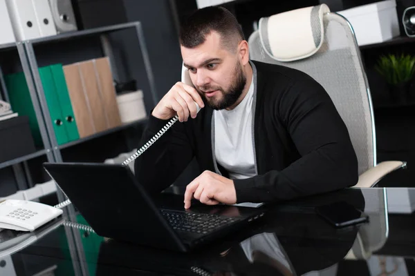 Busy young man talking on phone and looking at laptop in office — Stock Photo, Image