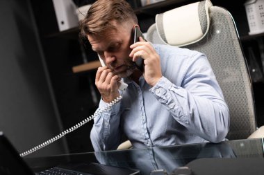 Resepsiyon bilgisayar başında oturan ve sabit telefon ve cep telefonu tutan genç adam