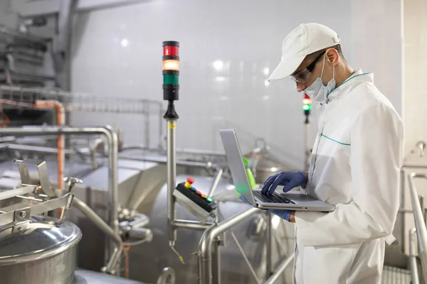 Homem em um manto branco e uma máscara com um laptop em suas mãos está na fábrica — Fotografia de Stock