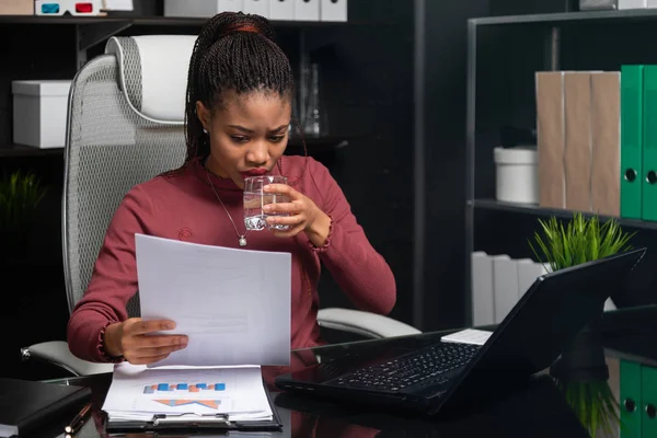 Hermosa joven mujer de negocios afroamericana sosteniendo un vaso de agua y trabajando con documentos en la mesa en la oficina — Foto de Stock