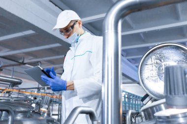 Beyaz bir elbise, eldiven ve maske bir tablet ve standları en sonda fabrika süt ürünleri tutarak teknoloji uzmanı. Müfettiş üretim tesislerinde performansını denetler