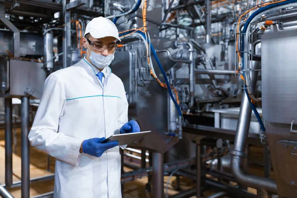 Man in een masker en een badjas stands met een tablet in zijn handen in de zuivel fabriek — Stockfoto