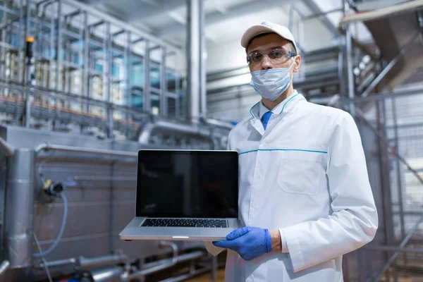 Uomo in vestaglia bianca e una maschera con un computer portatile in mano è in fabbrica — Foto Stock