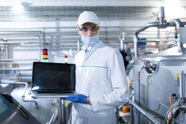 Homem em um manto branco e uma máscara com um laptop em suas mãos está na fábrica — Fotografia de Stock