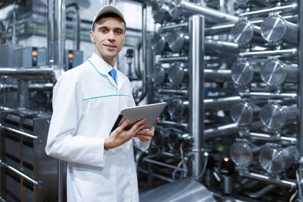 Homem em um manto branco com um tablet em suas mãos está na loja de produção na fábrica — Fotografia de Stock