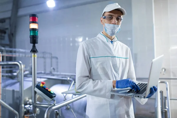 Uomo in vestaglia bianca e una maschera con un computer portatile in mano è in fabbrica — Foto Stock