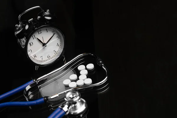 Stetoscopio e pillole sono accanto alla sveglia sul tavolo nero — Foto Stock