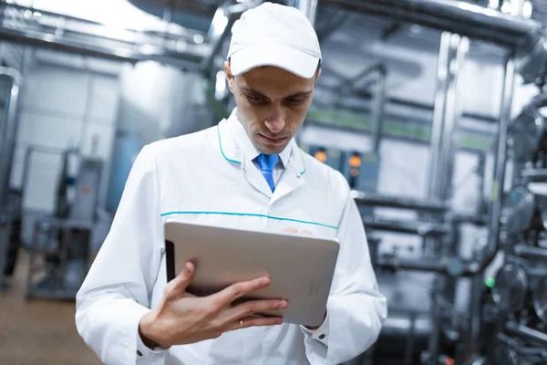 Retrato do homem com um manto branco e uma tampa em pé no departamento de produção da fábrica de laticínios com tablet cinza — Fotografia de Stock