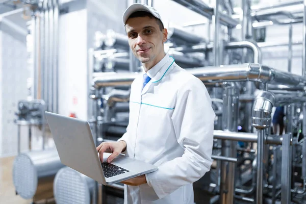 Retrato del hombre con una bata blanca y una gorra de pie en el departamento de producción de la fábrica de lácteos con ordenador portátil —  Fotos de Stock