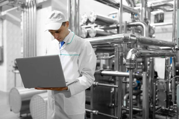 Retrato del hombre con una bata blanca y una gorra de pie en el departamento de producción de la fábrica de lácteos con ordenador portátil —  Fotos de Stock