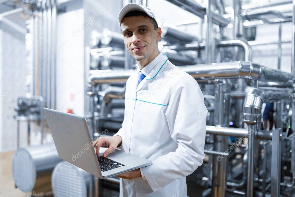 Portrait of man in a white robe and a cap standing in production department of dairy factory with laptop