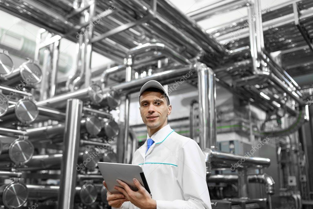 Portrait of man in a white robe and a cap standing in production department of dairy factory with grey tablet