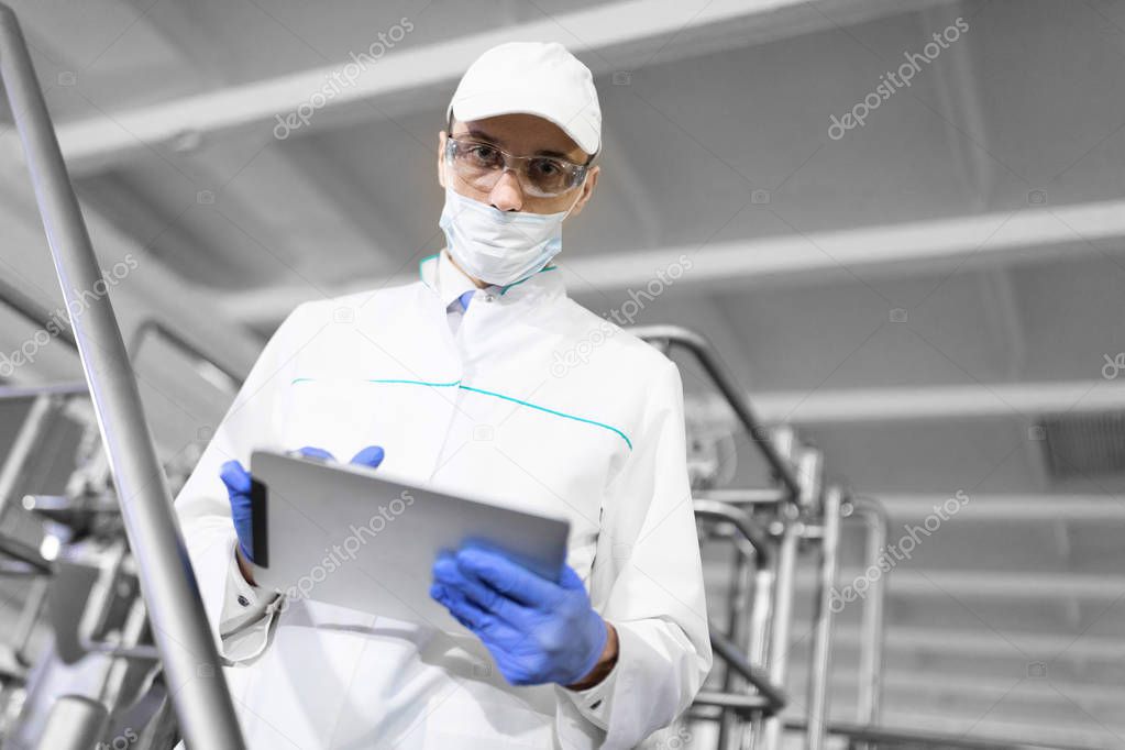 Portrait of man in a white robe and a cap standing in production department of dairy factory with grey tablet