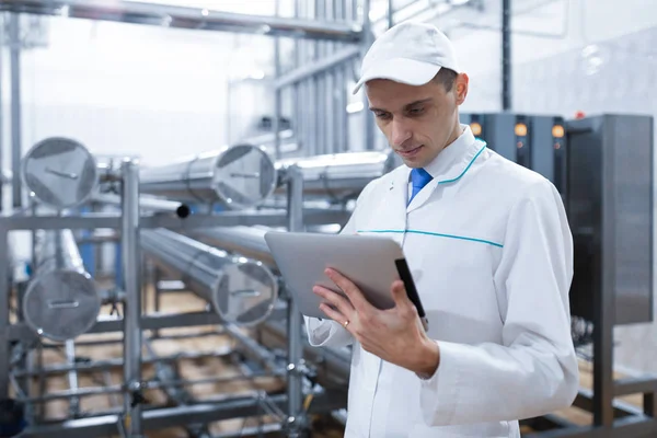 Retrato do homem com um manto branco e uma tampa em pé no departamento de produção da fábrica de laticínios com tablet cinza — Fotografia de Stock