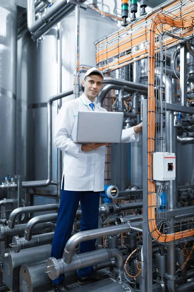 Retrato del hombre con una bata blanca y una gorra de pie en el departamento de producción de la fábrica de lácteos con ordenador portátil —  Fotos de Stock