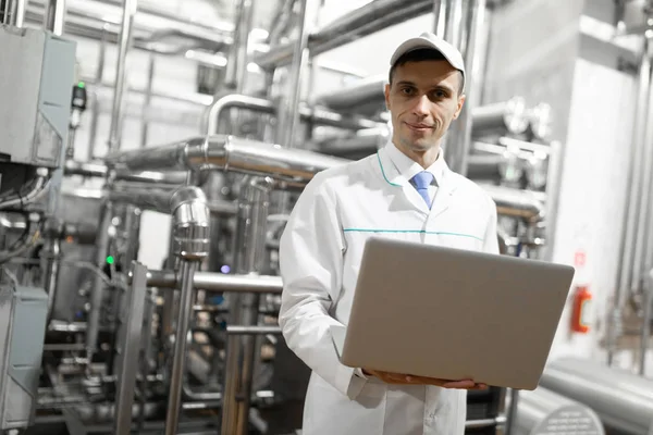 Retrato de homem em um manto branco e um boné em pé no departamento de produção de fábrica de laticínios com laptop — Fotografia de Stock