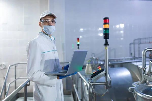 Retrato del hombre con una bata blanca y una gorra de pie en el departamento de producción de la fábrica de lácteos con ordenador portátil — Foto de Stock