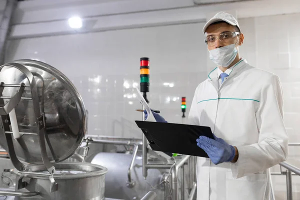Инспектор в маске и скрабе стоит с папкой-планшетом в руках на молочном заводе — стоковое фото